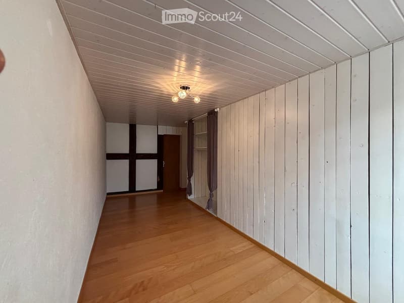 Empty hallway with wooden flooring, white paneled walls, and a slanted white ceiling with recessed lighting. A door and a window with curtains are visible at the end of the hallway.