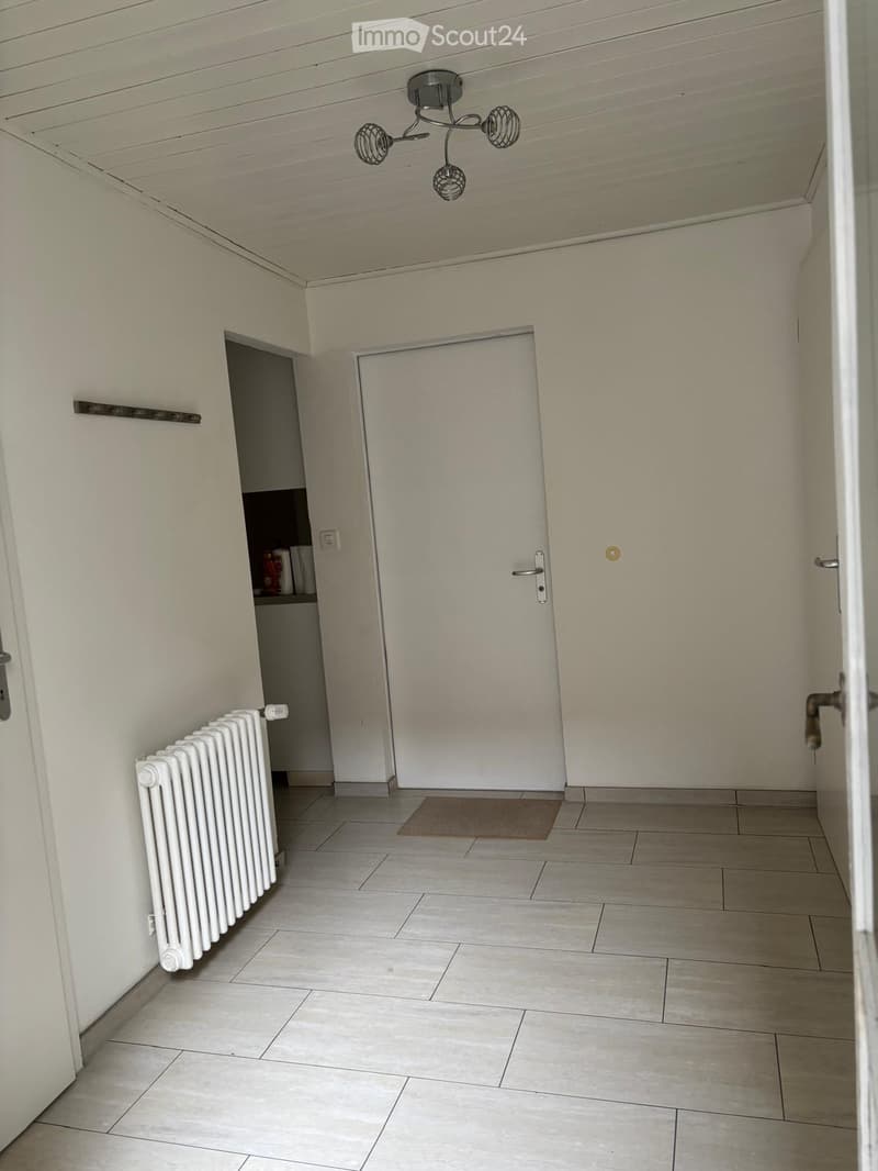 Empty hallway with tiled floor, white walls, radiator, and a small shelf with items visible in the background.