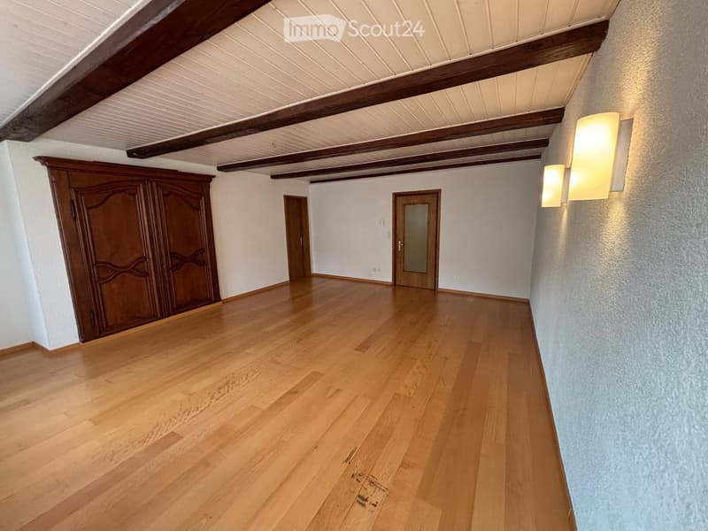 Empty room with wooden flooring, white walls, exposed wooden beams on the ceiling, and two doors, one with a glass panel. Wall-mounted light fixtures are visible on the right side.