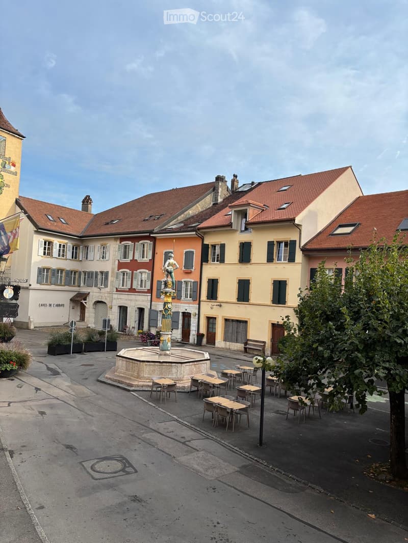 Courtyard with a central fountain, surrounded by tables and chairs, with several buildings featuring red-tiled roofs and shutters.