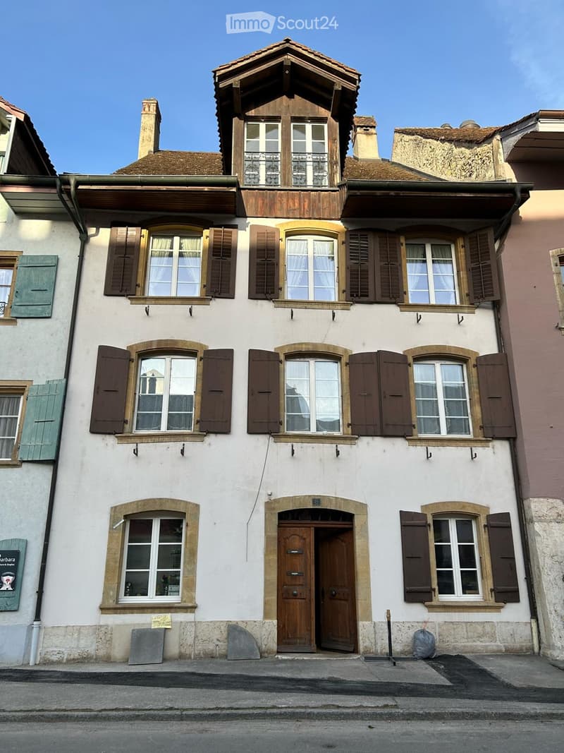 A three-story residential building with white walls, multiple windows with shutters, and a wooden front door. The roof features a small attic with dormer windows and a balcony.