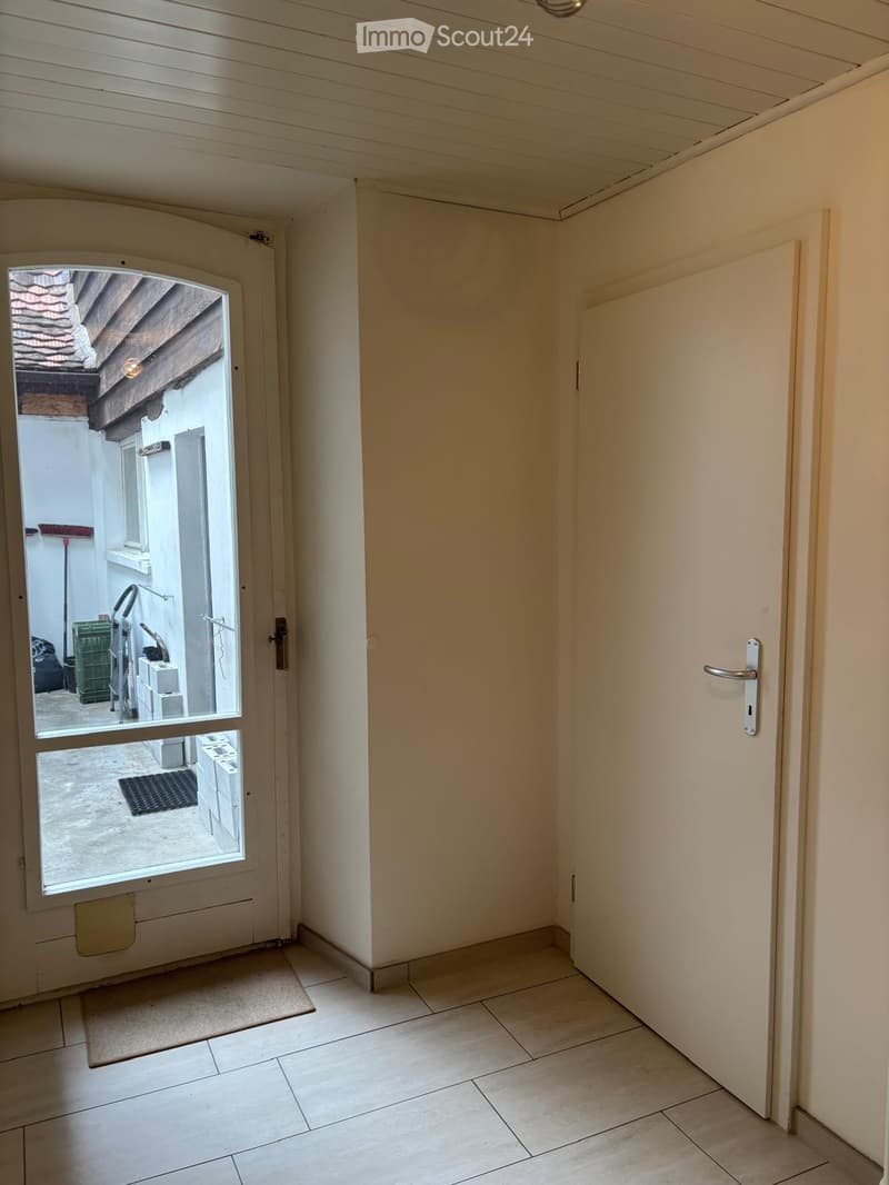 Empty hallway with tiled floor, white walls, and a door leading to an outdoor area. The outdoor area includes a small mat, a broom, and some stacked items.