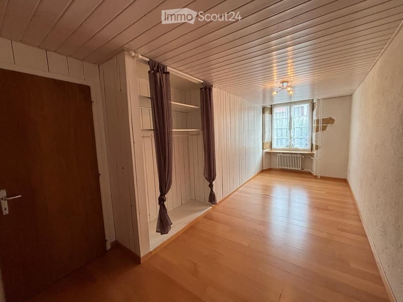 Empty room with wooden flooring, white paneled walls, and a large window with curtains. The room features a built-in closet with shelves and a radiator under the window.