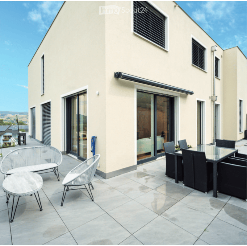 Modern outdoor terrace with a glass-walled building in the background, featuring a white wicker sofa, a round glass coffee table, and a black dining set with chairs.