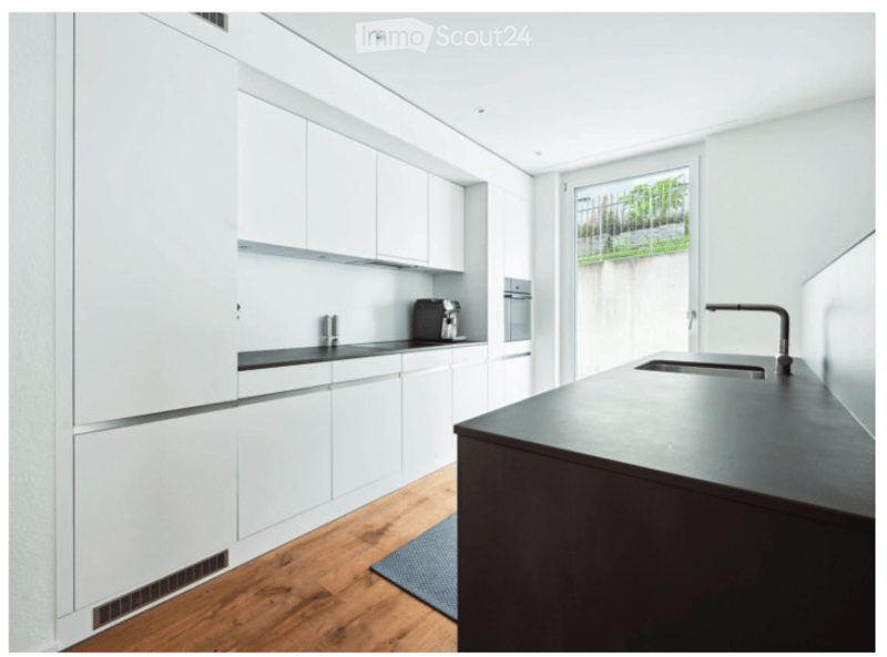 Modern kitchen with white cabinets, dark countertop, built-in oven, and a sink with a modern faucet. The space features wooden flooring and a small rug near the island.