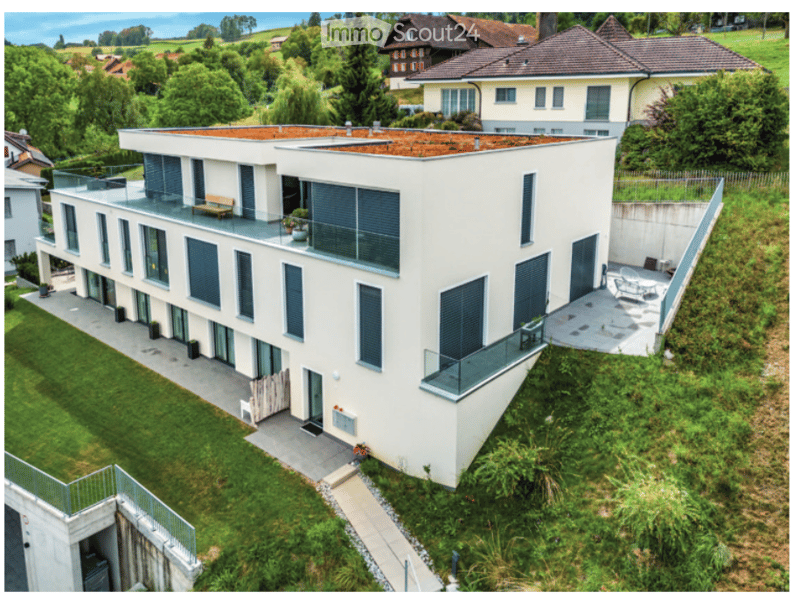 Modern two-story house with white exterior, large windows, balconies, and a flat roof. The house is surrounded by a well-maintained garden with greenery and a pathway leading to the entrance.