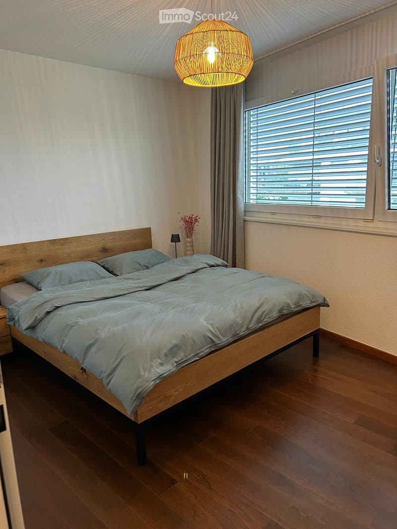 Spacious bedroom with wooden bed frame, gray bedding, and a woven pendant light fixture. Large window with blinds provides natural light and a view.