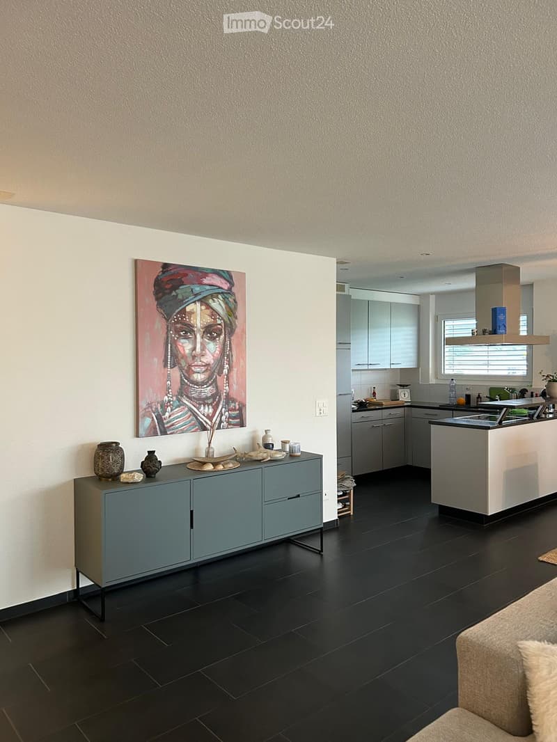 {"visible_features": ["Fireplace"],"type": "Living room and kitchen","description": "The image shows an open-concept living room and kitchen area. The living room features a large painting of a woman's face on the wall, with a gray sideboard underneath.