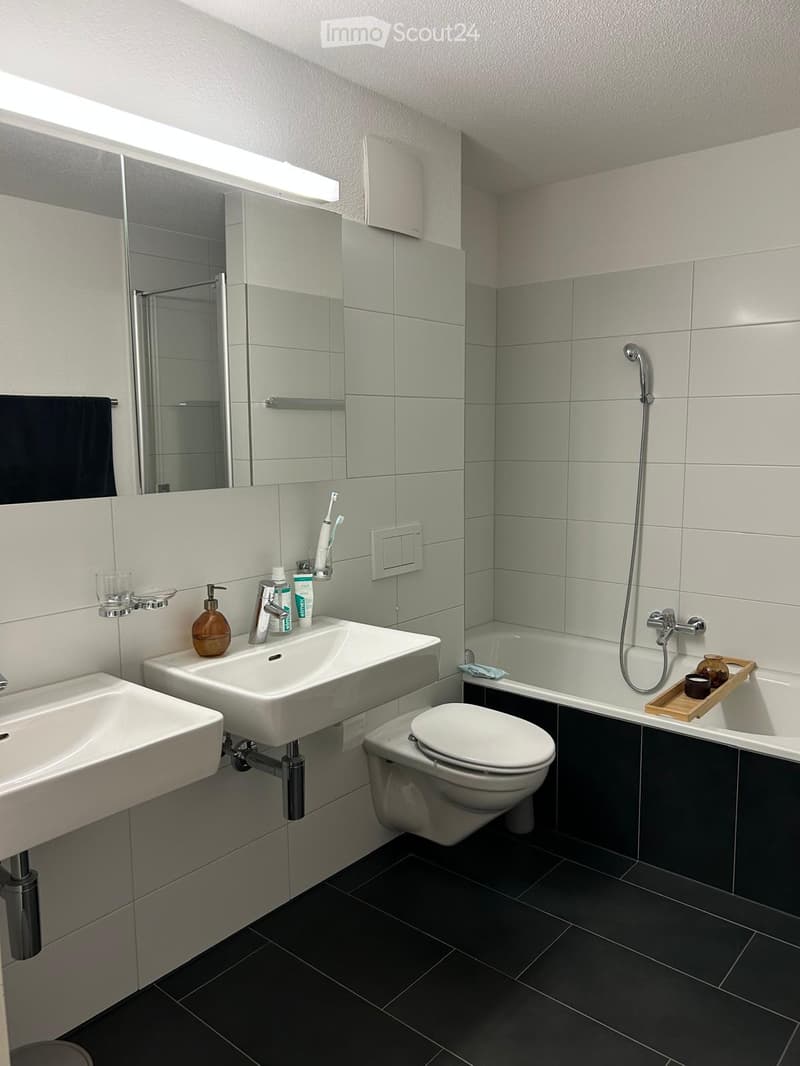 Modern bathroom with white tiles, double sink vanity, toilet, and shower area with glass enclosure
