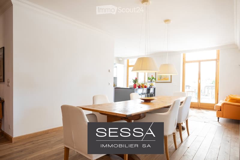 Bright, open-plan living and dining area with wooden flooring, white walls, large windows, a wooden dining table with chairs, a sofa, pendant lamps, and a small kitchen area with stools and plants.