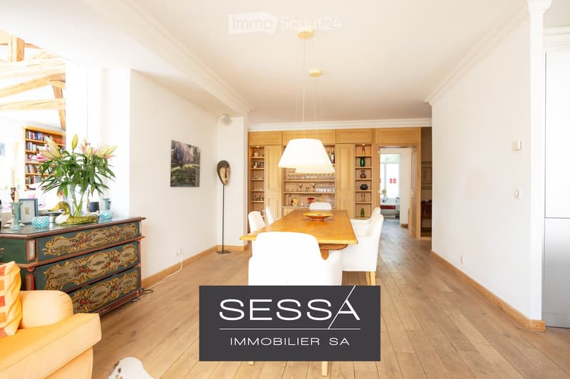 Bright, open living and dining area with wooden flooring, white walls, and large windows. Features a dining table with chairs, a sofa, a cabinet, and decorative items including a vase with flowers and a picture frame on the wall.