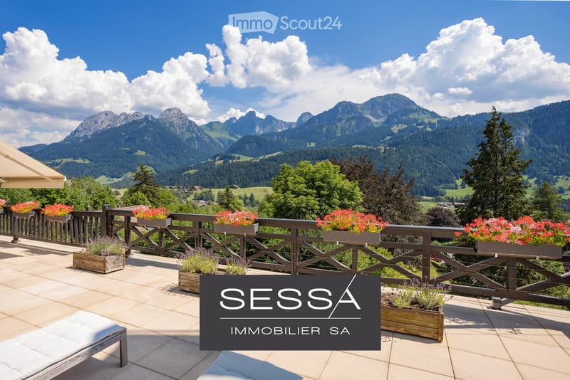 A spacious balcony with wooden planters filled with flowers and shrubs, overlooking a scenic mountain landscape with lush green valleys and scattered houses.