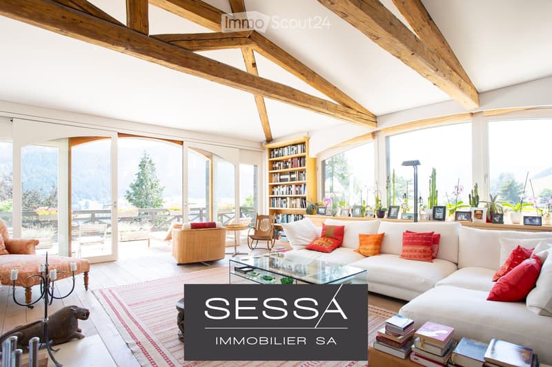 Bright living room with exposed wooden beams, large windows offering a scenic view, a white sectional sofa with colorful cushions, a glass coffee table, a wooden chair, a bookcase, and decorative items including a candle holder and a statue.