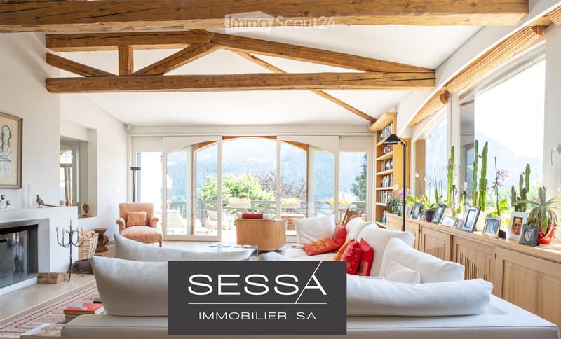 Bright living room with exposed wooden beams, large windows offering a scenic mountain view, a white sofa with red cushions, a coffee table, an armchair, a bookshelf, and a fireplace.
