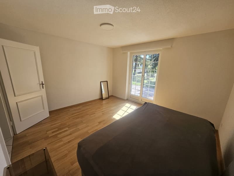 A well-lit bedroom with wooden flooring, a large bed with dark bedding, a glass table, a mirror, and a sliding glass door leading to a garden.