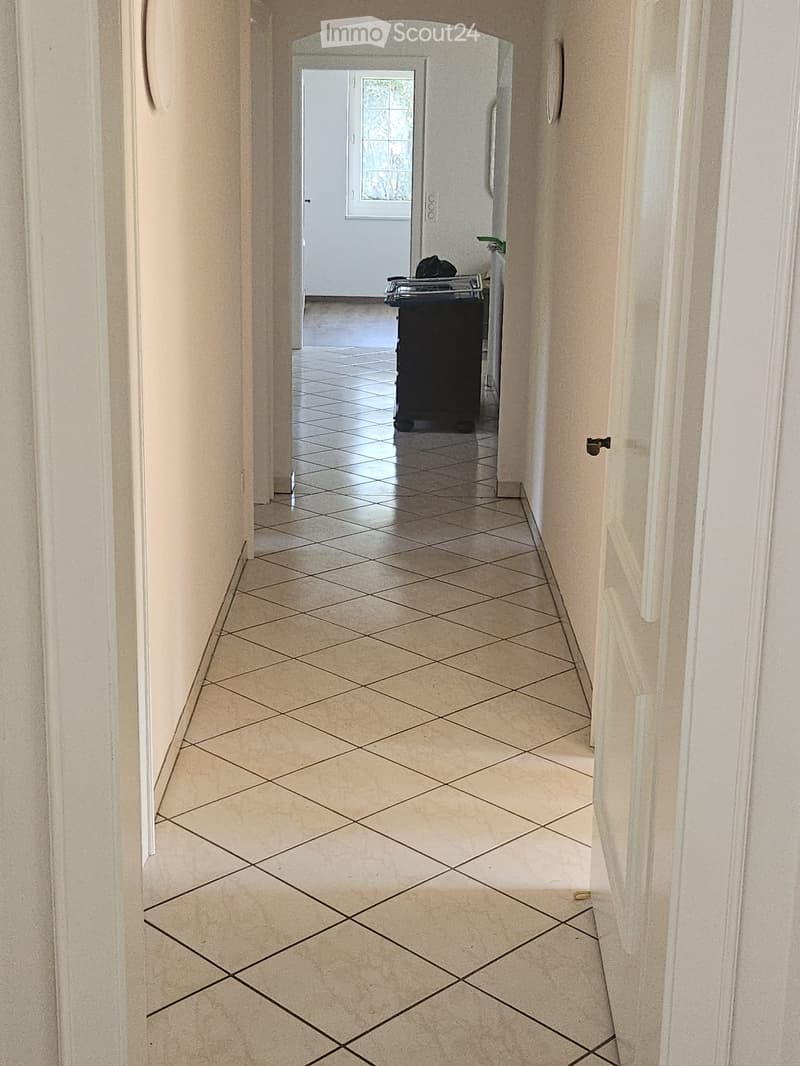 A clean, tiled hallway with beige walls and a patterned tile floor, leading to a room with a visible door and window.