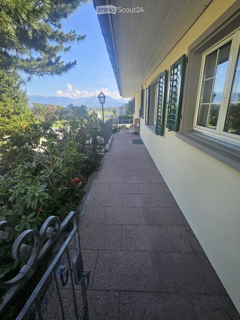 A paved pathway leading to a building entrance, flanked by greenery and a metal railing on one side.