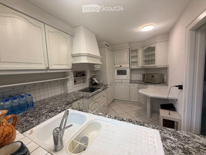 Modern kitchen with white cabinets, granite countertops, stainless steel sink, and appliances including an oven and microwave. Features a double sink with a faucet, water bottles on the counter, and a small stool near the corner.