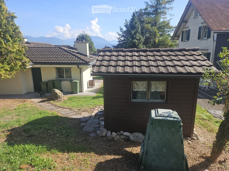 A small wooden shed with a tiled roof is situated in a garden area with grass, stone pathways, and surrounding trees. Nearby, there are additional buildings and a view of mountains in the background.