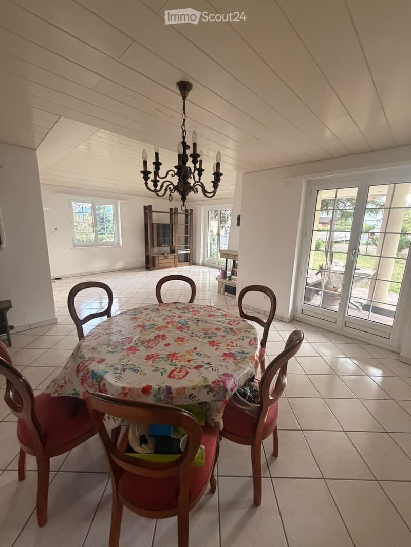 Bright dining room with a round table covered in a floral tablecloth, surrounded by four wooden chairs with red cushions. A chandelier hangs from the sloped ceiling, and a shelf with a television is visible in the background. Large windows and glass doors