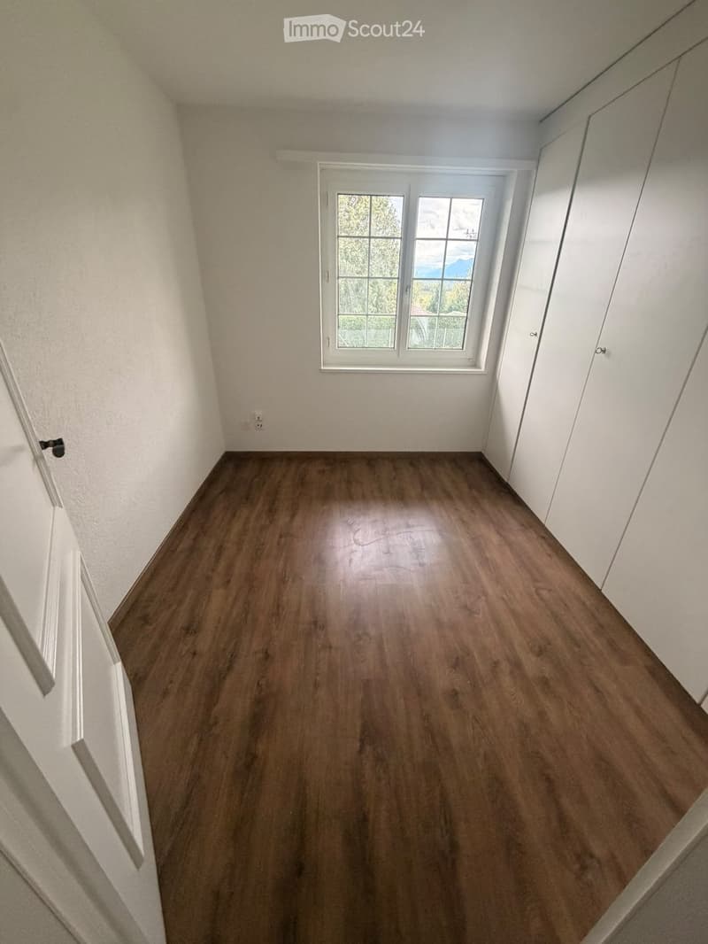Empty room with wooden flooring, white walls, and a large window with a view of greenery outside.
