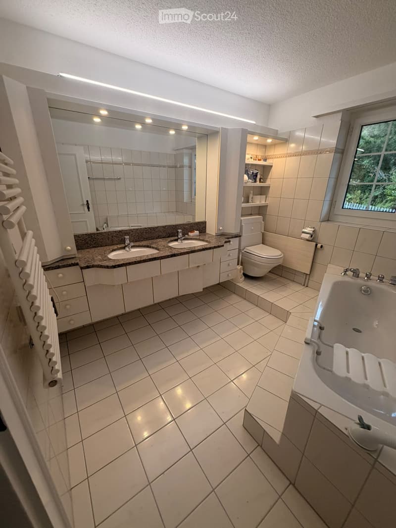 Modern bathroom with tiled floors and walls, double sink vanity, bathtub, toilet, large mirror, and built-in cabinet with shelves.
