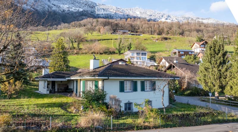 Single-story house with green shutters and a tiled roof, situated in a scenic valley with snow-capped mountains in the background.