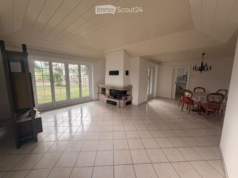 Bright living room with tiled floor, large windows, fireplace, dining area with table and chairs, and chandelier.