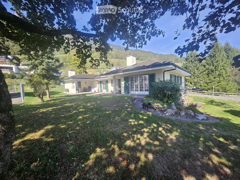 A two-story house with a tiled roof, green shutters, and large windows, surrounded by a well-maintained lawn and trees, with a pathway leading to the entrance.