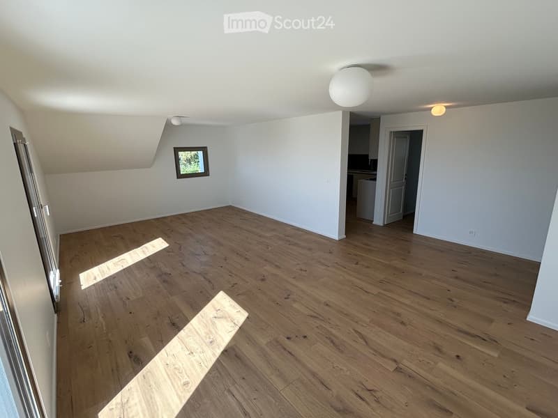 Empty room with wooden flooring, white walls, and a small window. The room has a slanted ceiling and a doorway leading to another area.
