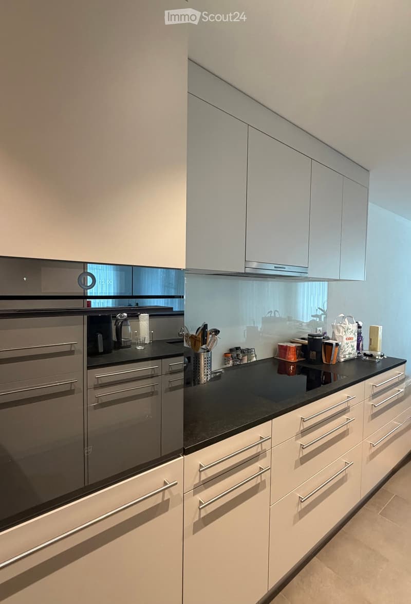 Modern kitchen with light-colored cabinets, stainless steel appliances, and a black countertop. Coffee maker, utensils, and various items are placed on the counter.
