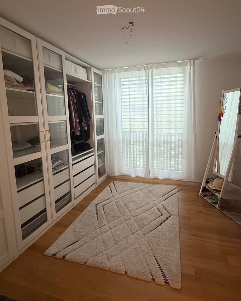 A well-lit bedroom with a large white wardrobe, a soft white rug, and a full-length mirror. The wardrobe contains folded clothes and shoes, and a hat is placed near the mirror.