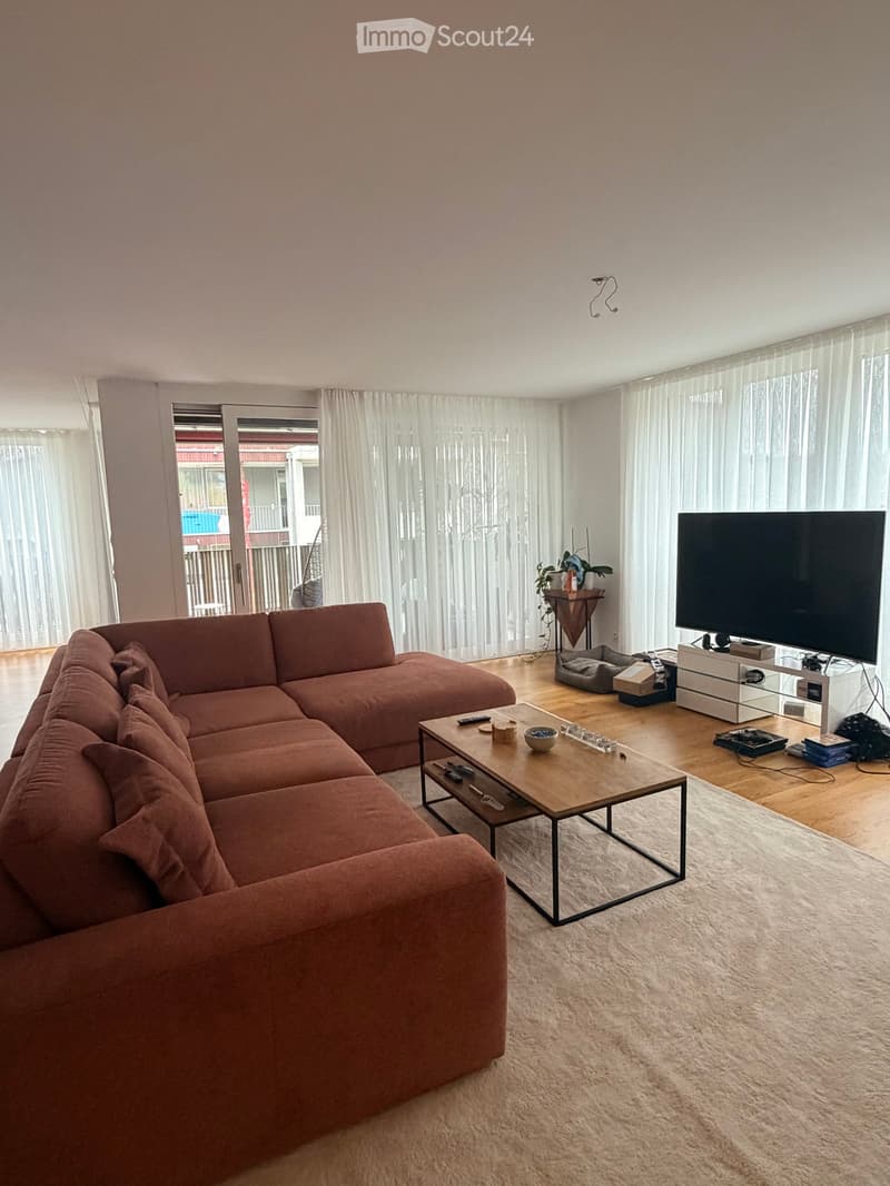 Modern living room with a large sectional sofa, coffee table, TV, and a small table with a plant. Large windows with sheer curtains and access to a balcony.