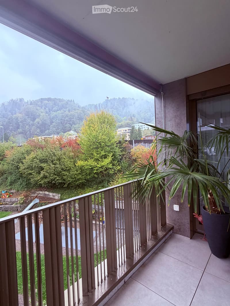 Balcony with a metal railing, tiled floor, and a large potted plant, overlooking a green, hilly landscape.