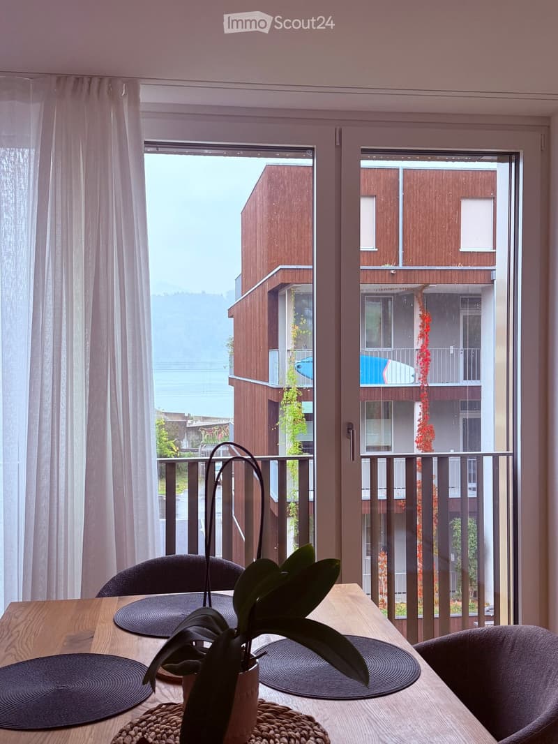 Dining area with a wooden table, black placemats, and a potted plant, overlooking a modern building exterior with balconies and greenery.
