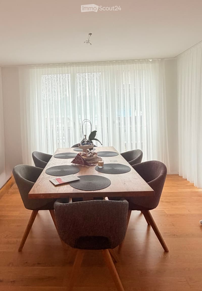 A modern dining room with a wooden dining table surrounded by six grey chairs, placemats, a plant, and a cup on the table. Large windows with sheer curtains allow natural light to fill the space.