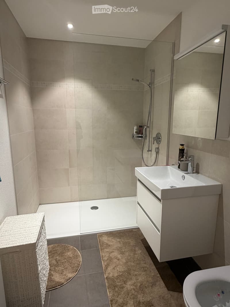 Modern bathroom with a bathtub, sink, toilet, shower, and a built-in cabinet. Features a beige tile design and a wicker basket on the side.