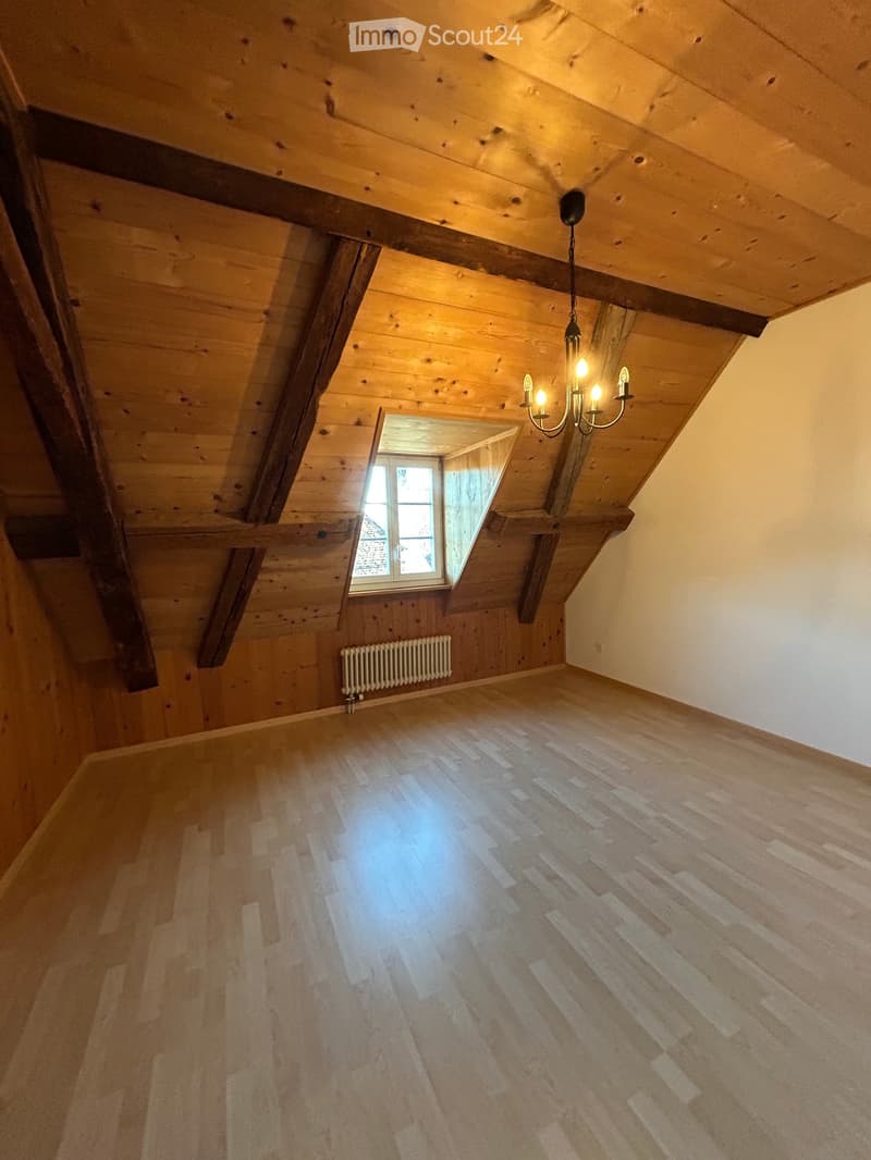 Empty attic room with wooden walls, exposed wooden beams, wooden floor, skylight window, radiator, and chandelier light fixture.