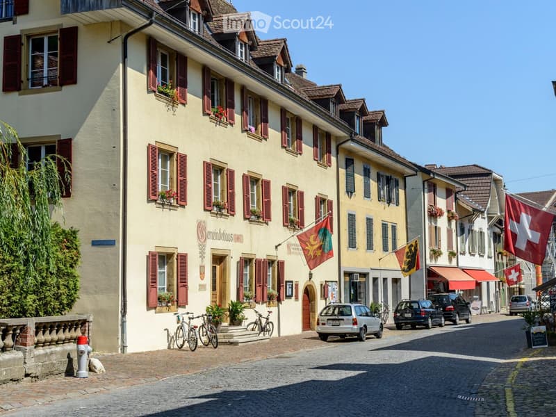 A charming European street lined with multi-story buildings featuring red shutters, flower boxes, and Swiss flags. The street is paved with cobblestones and has parked cars and bicycles.