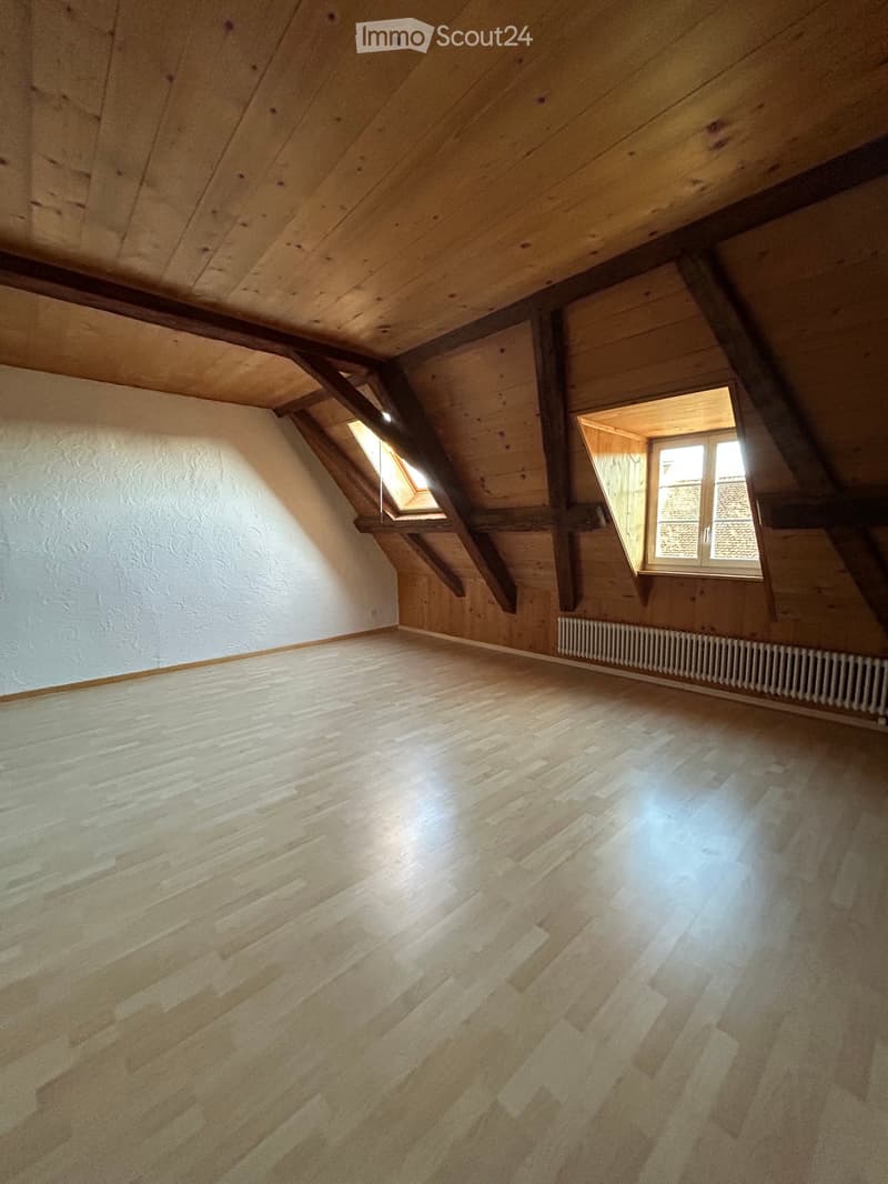 Empty attic room with wooden floors, exposed wooden beams, white walls, and two windows allowing natural light.