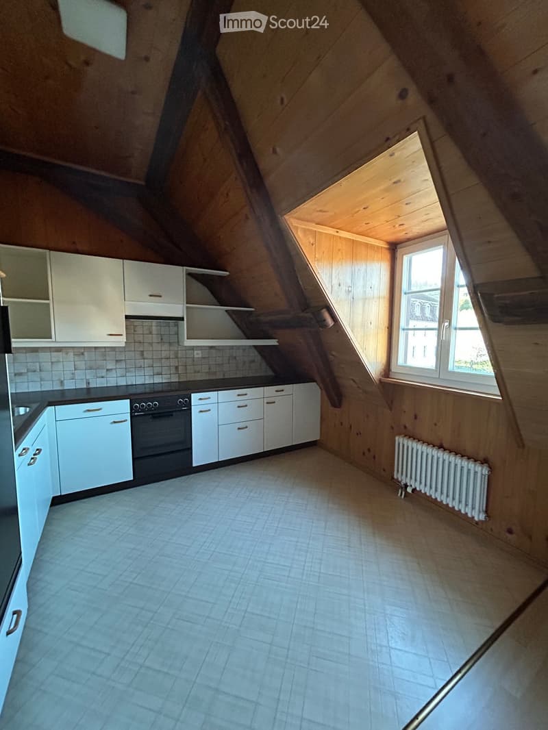 Attic kitchen with white cabinets, an oven, and a radiator, featuring wooden walls and a slanted ceiling with a window letting in natural light.