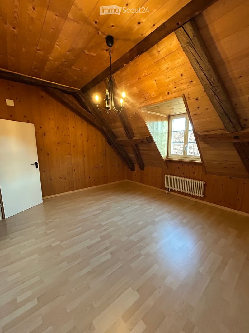Empty attic room with wooden walls and ceiling, exposed wooden beams, a chandelier, a window, and a radiator.