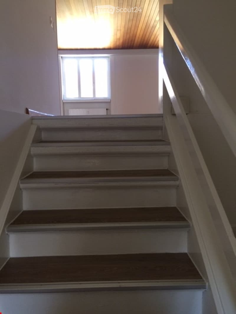 A narrow staircase with wooden steps and a white handrail, leading up to a landing with a window and a door at the top.