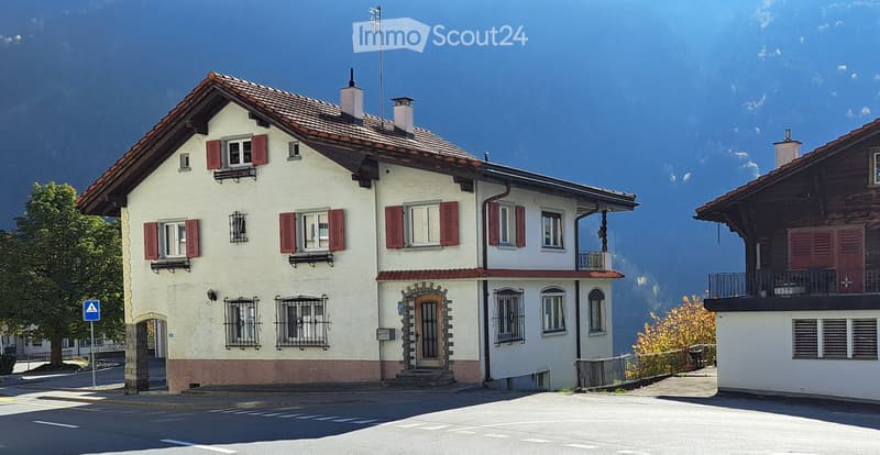 Traditional Swiss house with white walls, red shutters, and a tiled roof, situated in a scenic mountainous area.