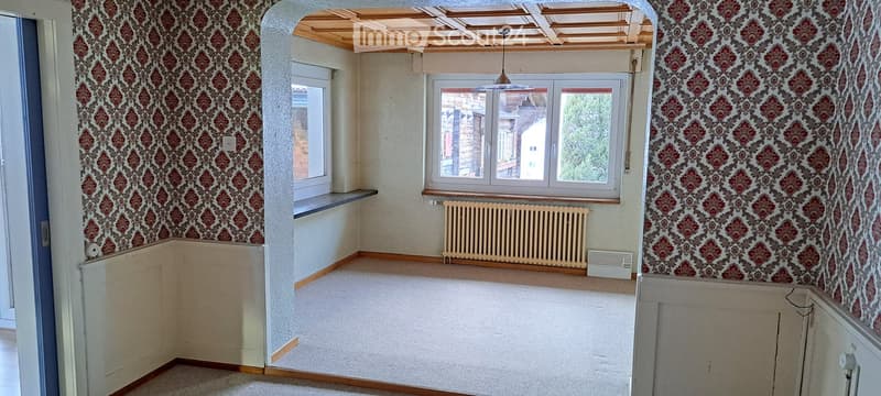 Empty room with patterned wallpaper, wooden floor, large windows, radiator, and exposed wooden ceiling beams.