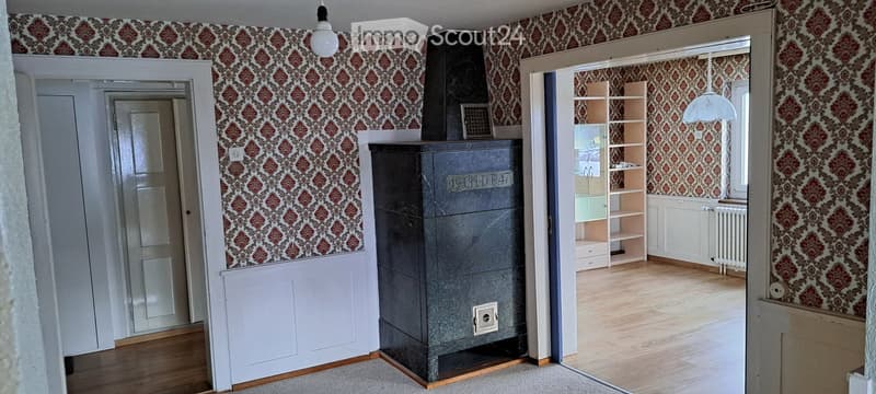 Empty room with wooden flooring, patterned wallpaper, and a large black stove in the center. A doorway leads to another room with a shelf unit and radiator.