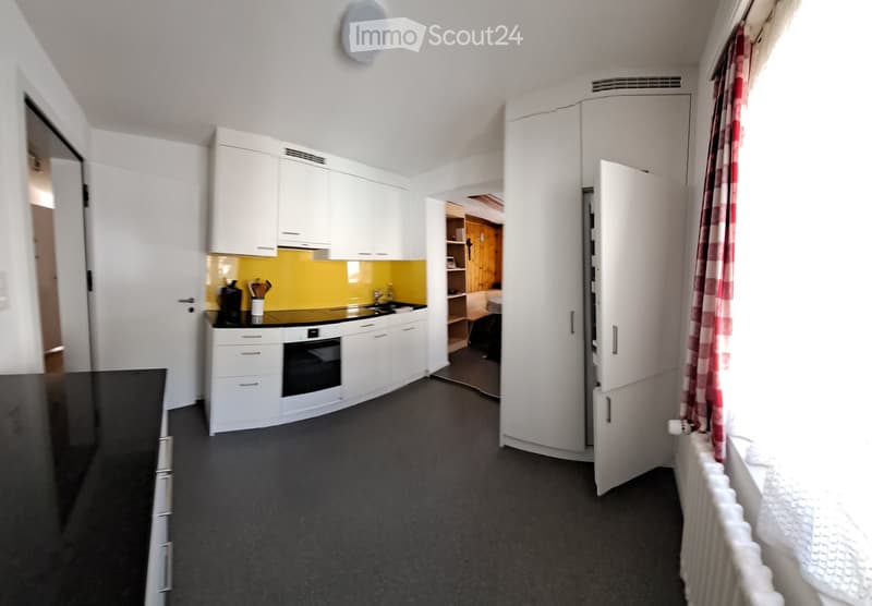 Modern kitchen with white cabinets, a black countertop, a sink, an oven, and a refrigerator. A red and white checkered curtain is visible on the window, and a radiator is placed below it.