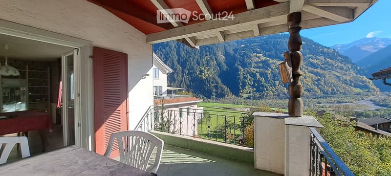 Balcony with a scenic mountain view, featuring a white plastic chair and a table, with a decorative wooden post and a view of the valley and surrounding houses.