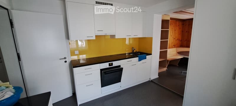 Modern kitchen with white cabinets, black countertop, stainless steel sink, and built-in oven. Adjacent room features wooden walls and a small table.