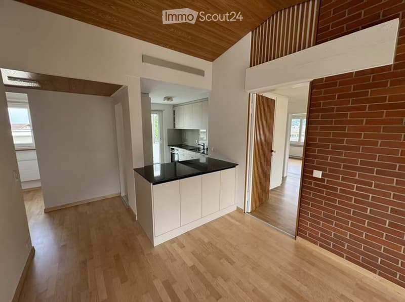 Open-plan kitchen area with white cabinets, black countertop, and a central island. Wooden flooring and exposed wooden ceiling beams.